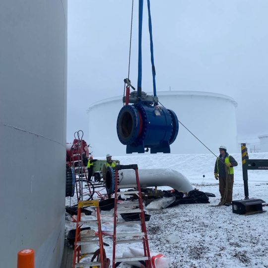 Workers crane-lifting large blue industrial valve at snowy facility